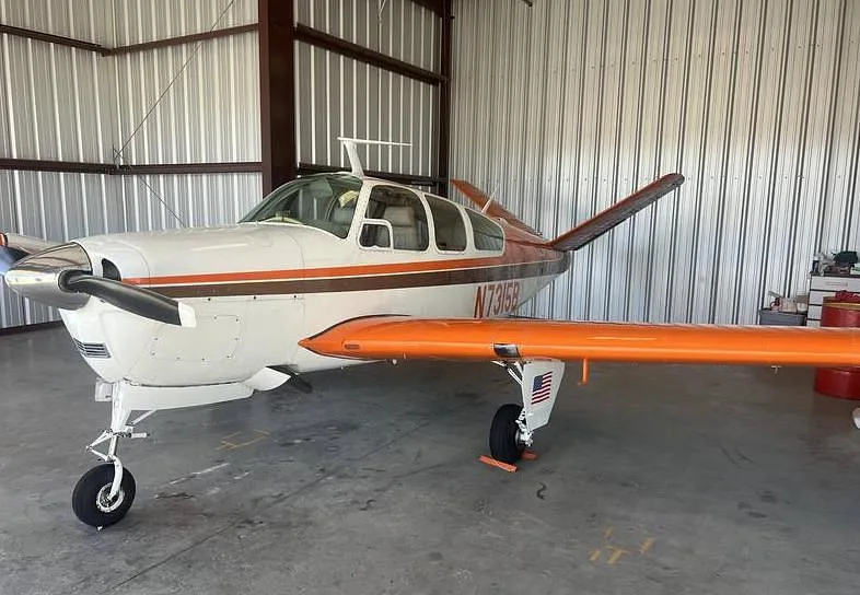A white and brown Beechcraft Bonanza N7315B with orange wings parked in a hangar, viewed from the front left.