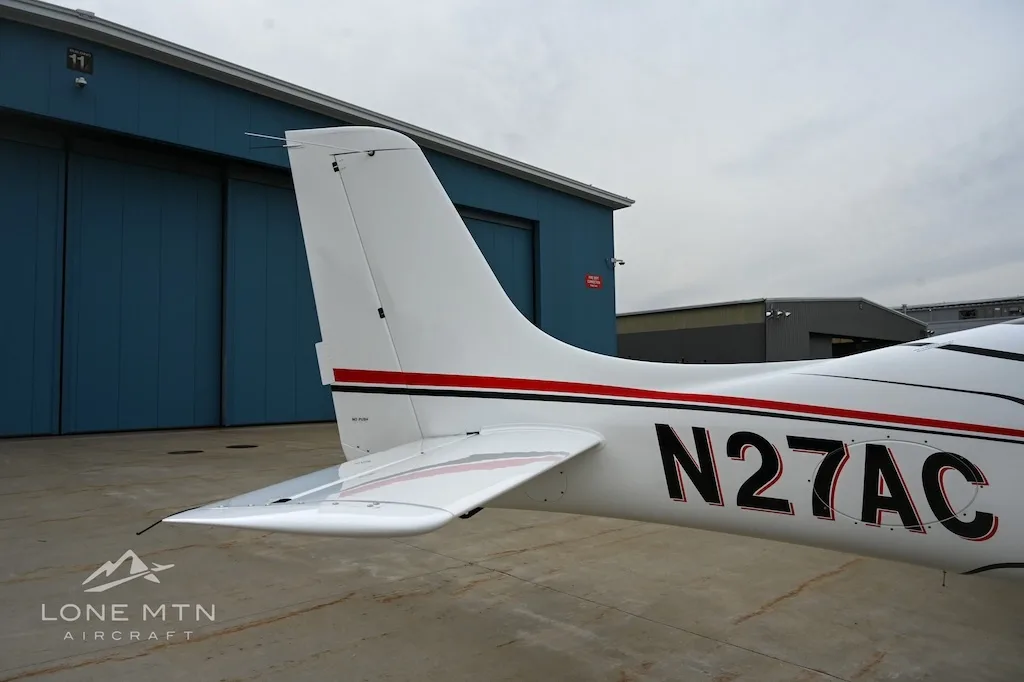 Close-up view of the tail section of a white Cirrus SR22 aircraft, tail number N27AC, against a blue hangar.