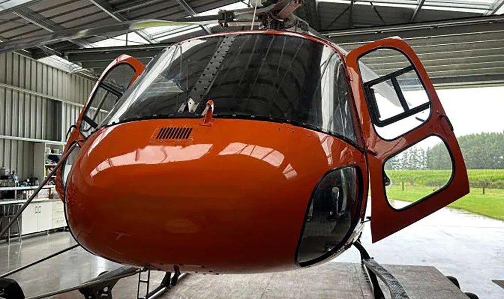A front view of a bright orange helicopter with both cockpit doors open, parked inside a hangar.