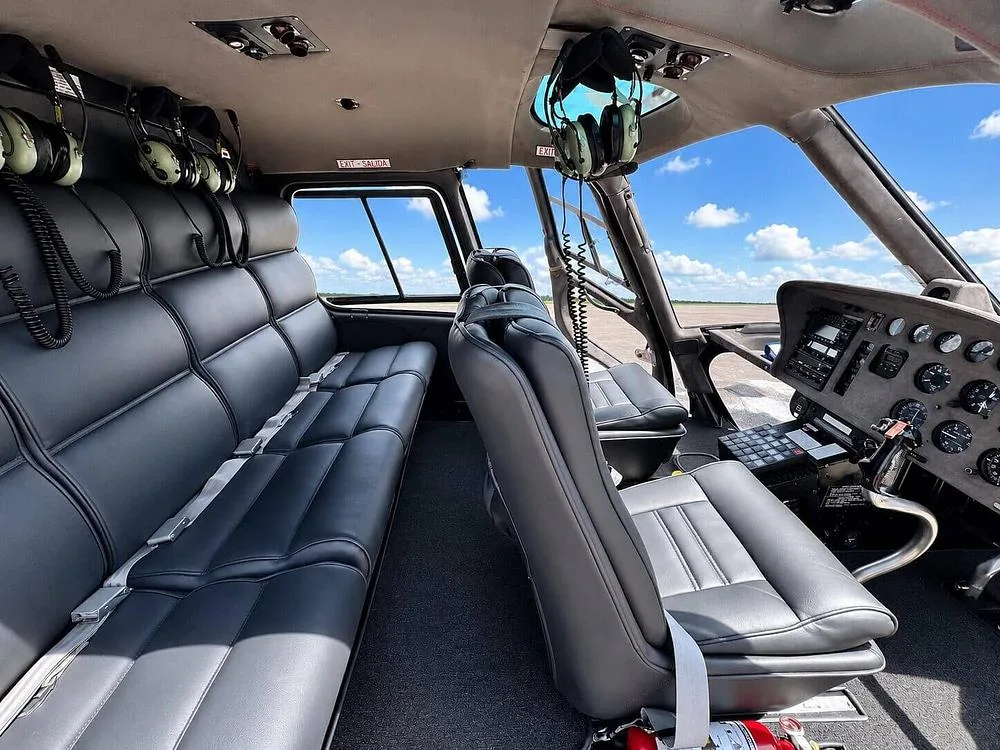 The interior of a helicopter cabin features black leather passenger seats and a partial view of the cockpit instruments.