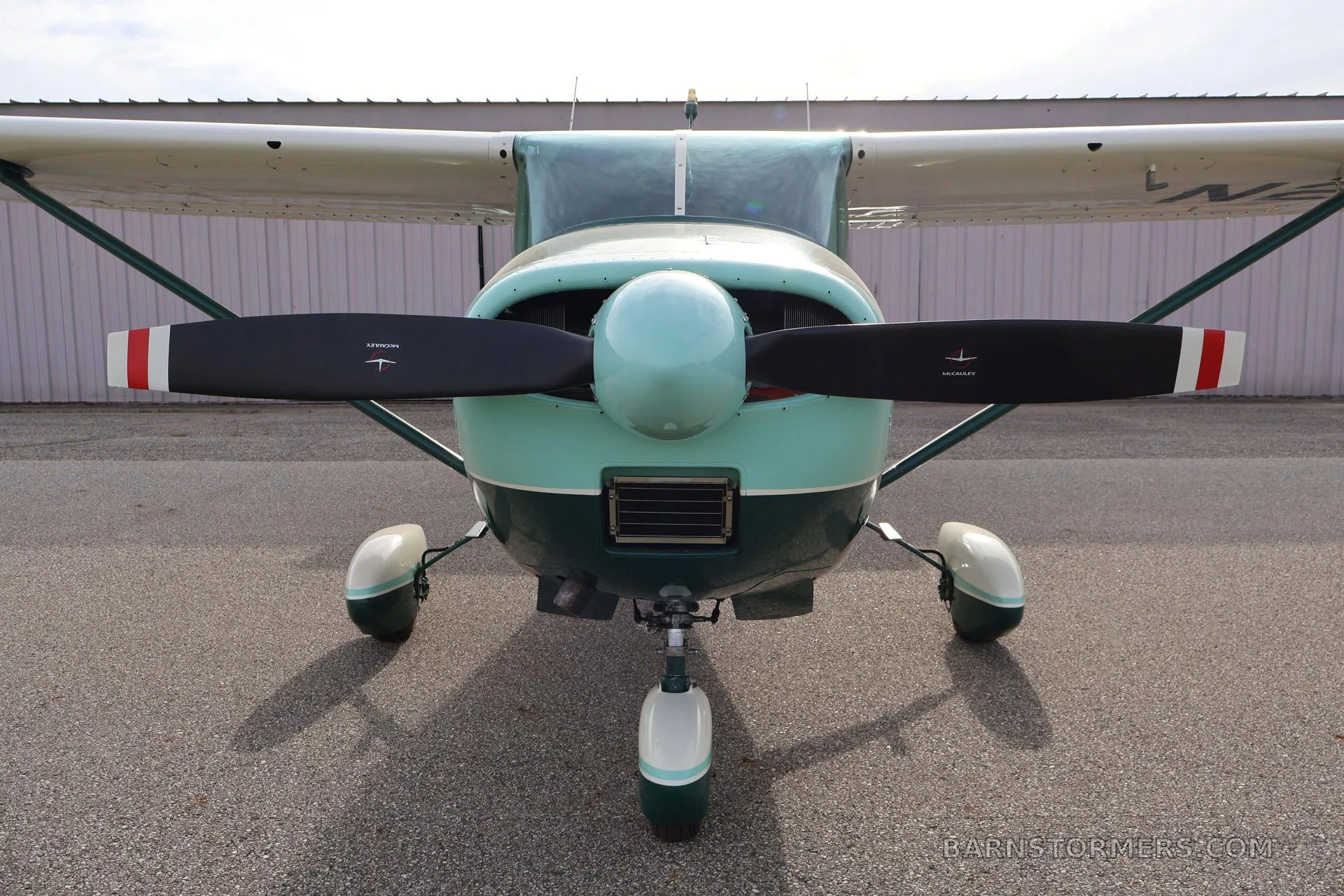 A green and white single-engine propeller aircraft (Cessna 172) is viewed head-on, showing its propeller and landing gear.