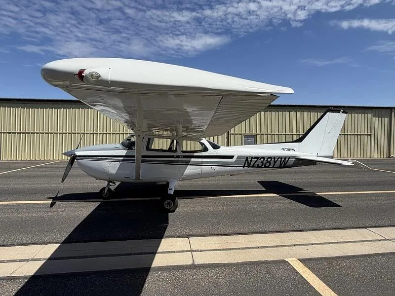 A side-left view of the white Cessna 172, N738YW, with gray and black stripes, parked on a tarmac next to a hangar.