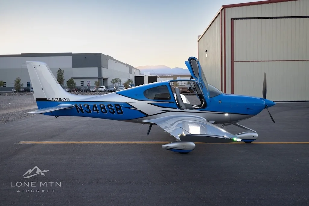 A side view of a blue and silver Cirrus SR22 aircraft with open doors parked on an airfield with buildings in the background.