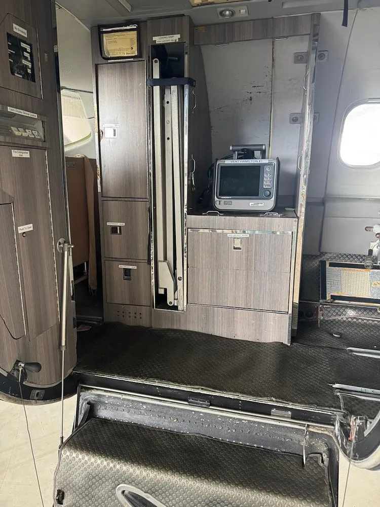 The interior of an aircraft cabin shows a galley area with wooden cabinets, a small monitor, and an open entry door with steps.