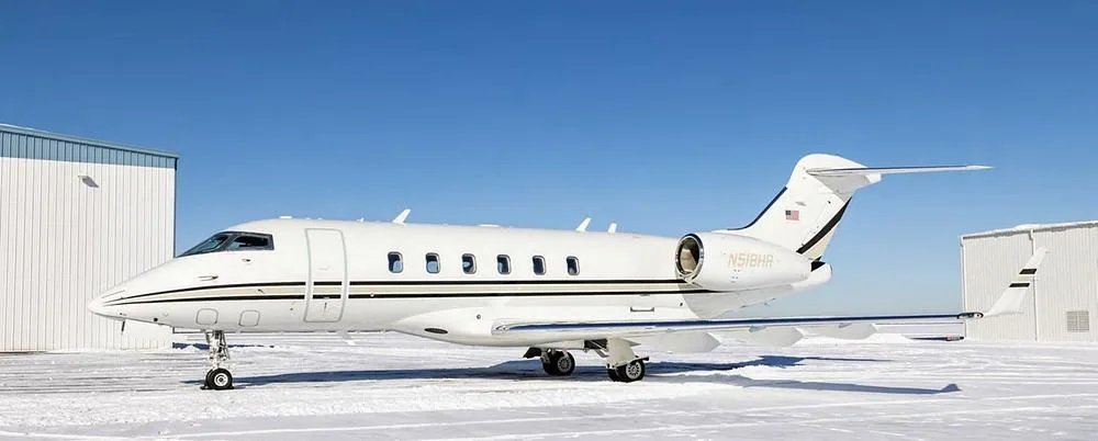 A white Bombardier Challenger business jet with black and gold stripes, tail number N518HR, parked on a snowy tarmac next to hangars.