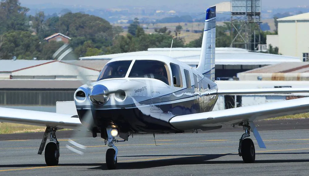 A white and blue Piper Saratoga II TC aircraft with its propeller spinning, on a taxiway.