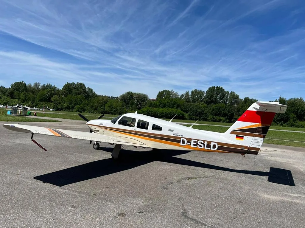 A side view of a white and brown Piper Arrow III (D-ESLD) parked on an airfield under a blue sky.