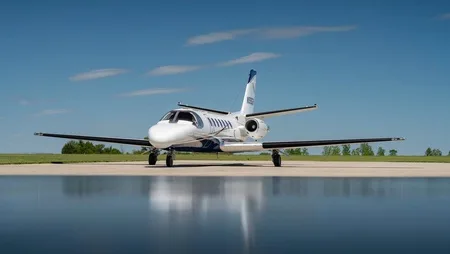 A white private jet with blue accents on the tail and belly, parked on an airfield under a clear blue sky, with a reflective surface in the foreground.