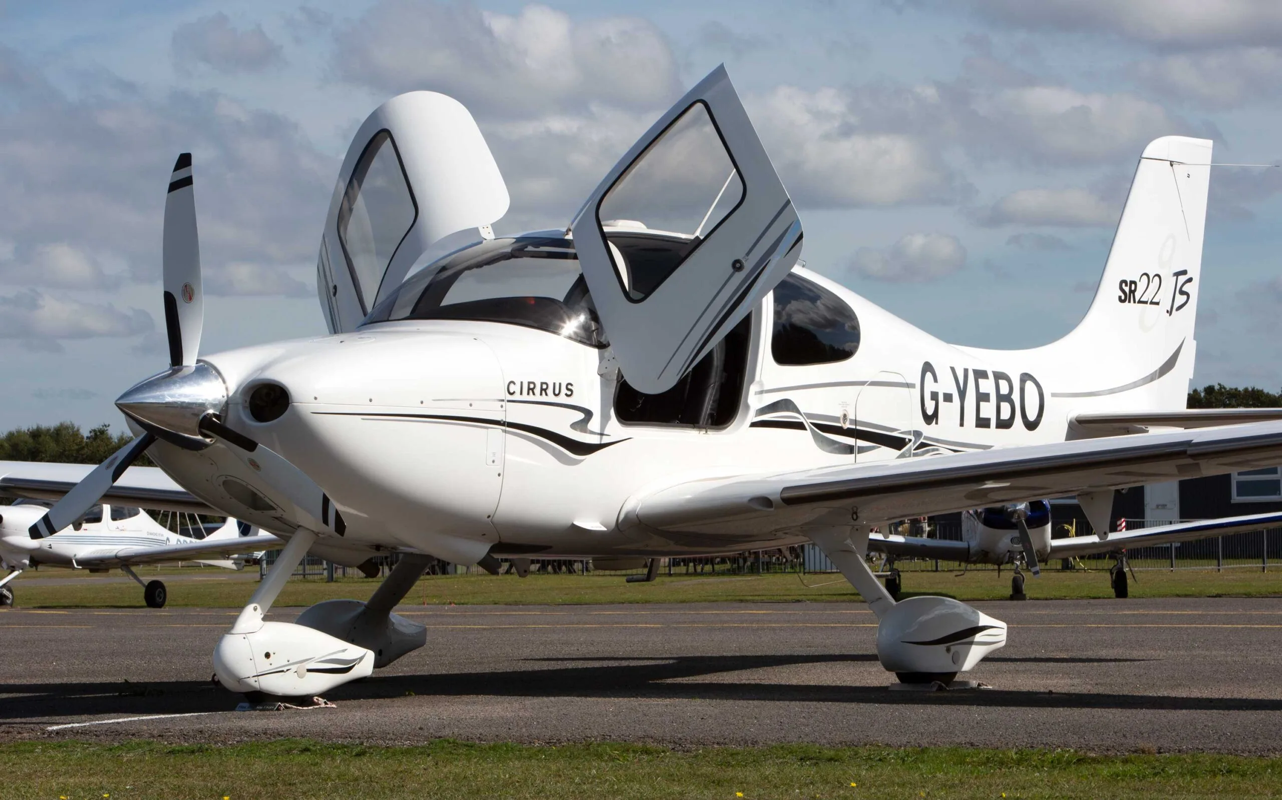 A white Cirrus SR22 TSi aircraft with black and grey stripes, registration G-YEBO, parked on an airfield with its gull-wing doors open.