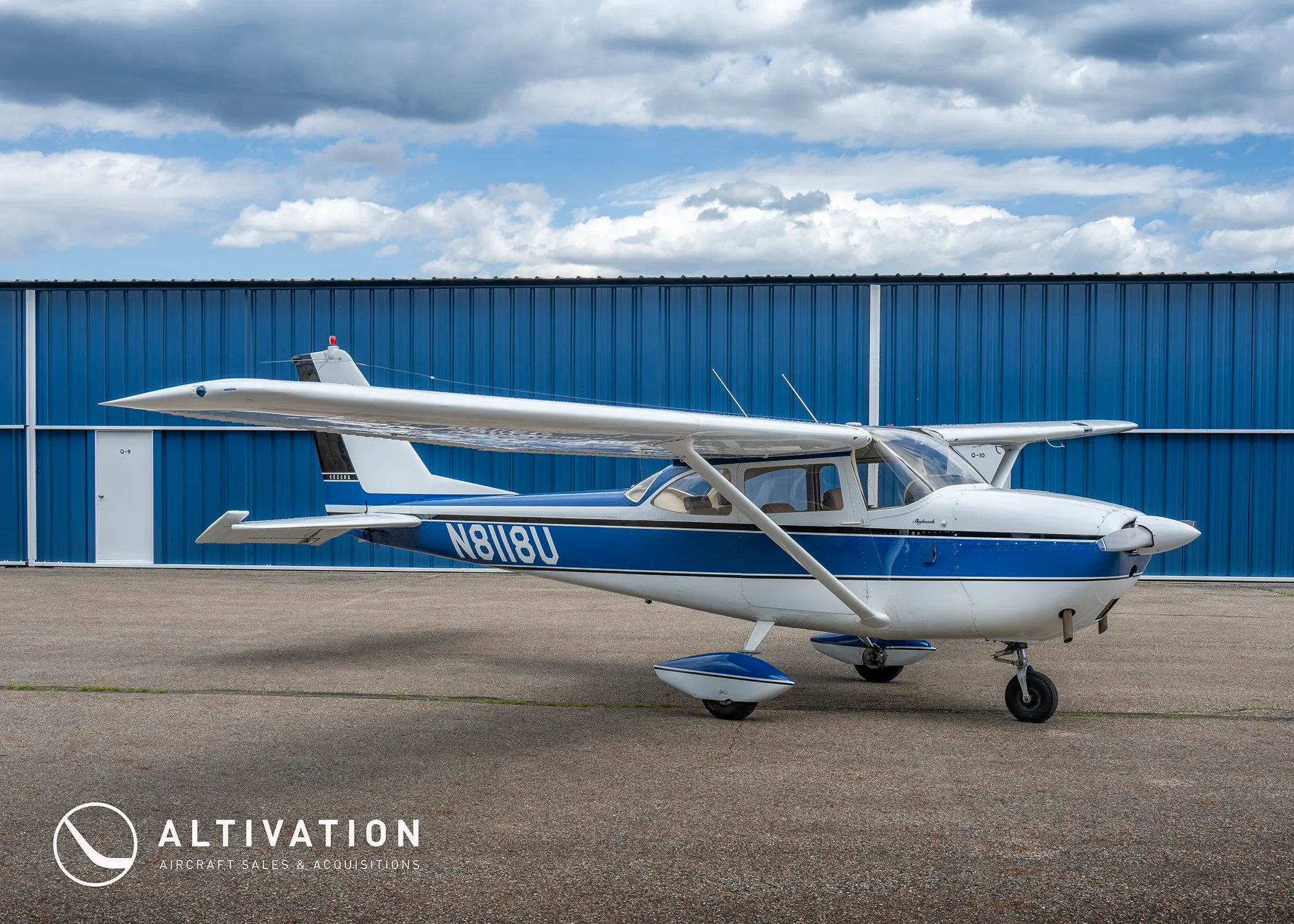 A side view of the white and blue Cessna 172 Skyhawk N8118U parked on an asphalt ramp in front of a blue hangar.