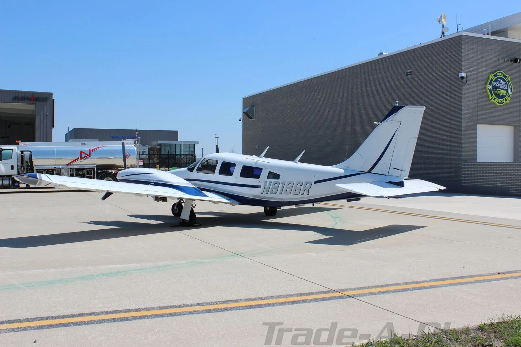 A side view of the white and blue Piper Seneca aircraft N8186R parked on a tarmac with a fuel truck in the background.