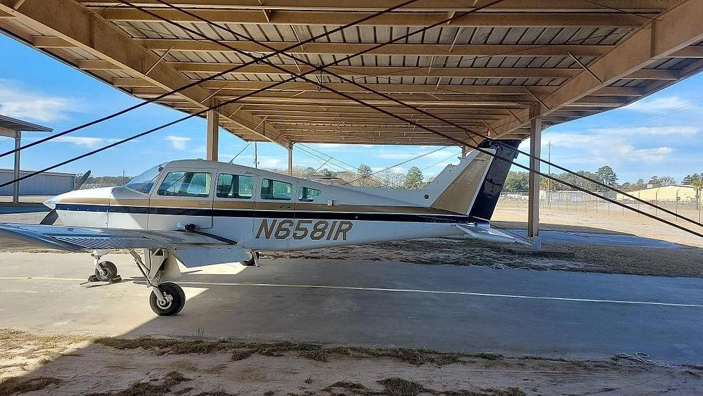 A white and gold single-engine propeller aircraft (Cessna 177 Cardinal) with registration N6581R is parked under an open-sided hangar, viewed from the side.