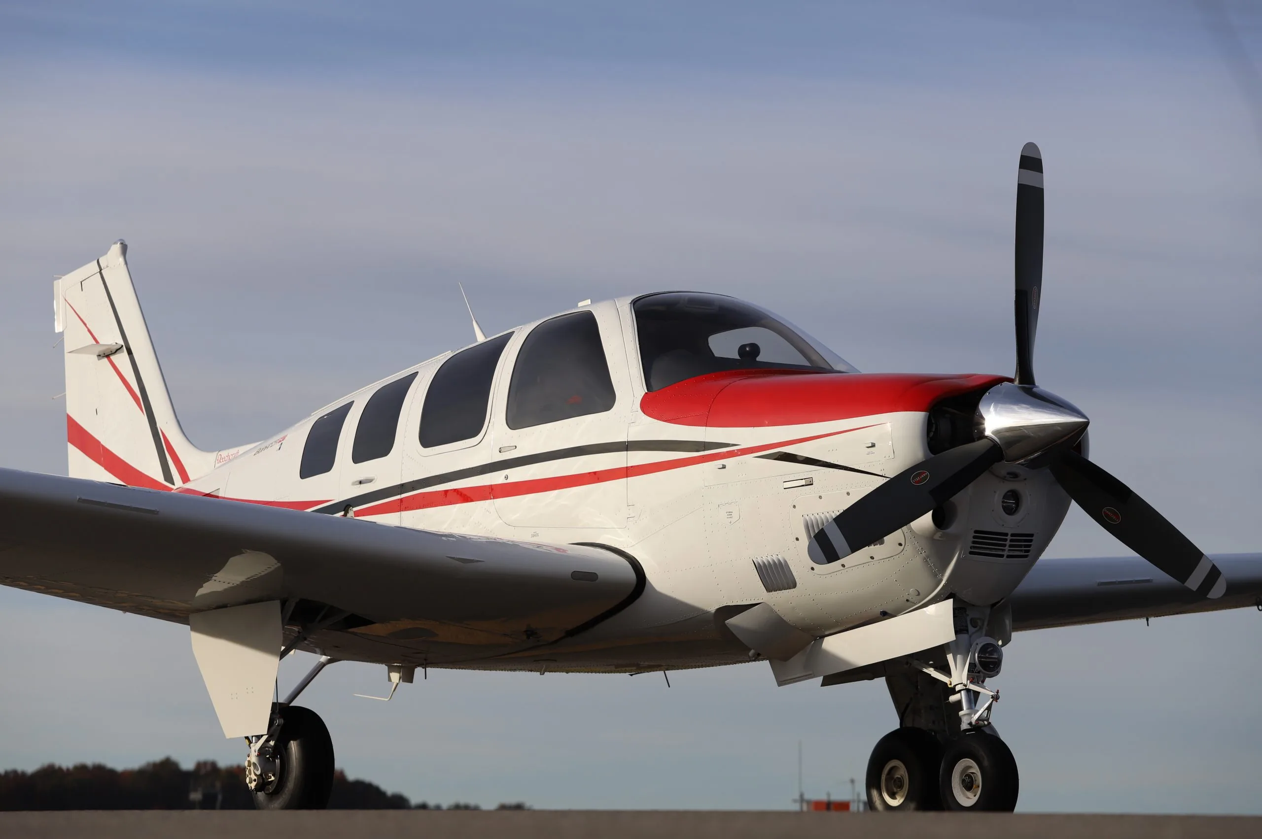 A white Beechcraft Bonanza with red and black stripes and a red nose cone sits on an airfield tarmac.