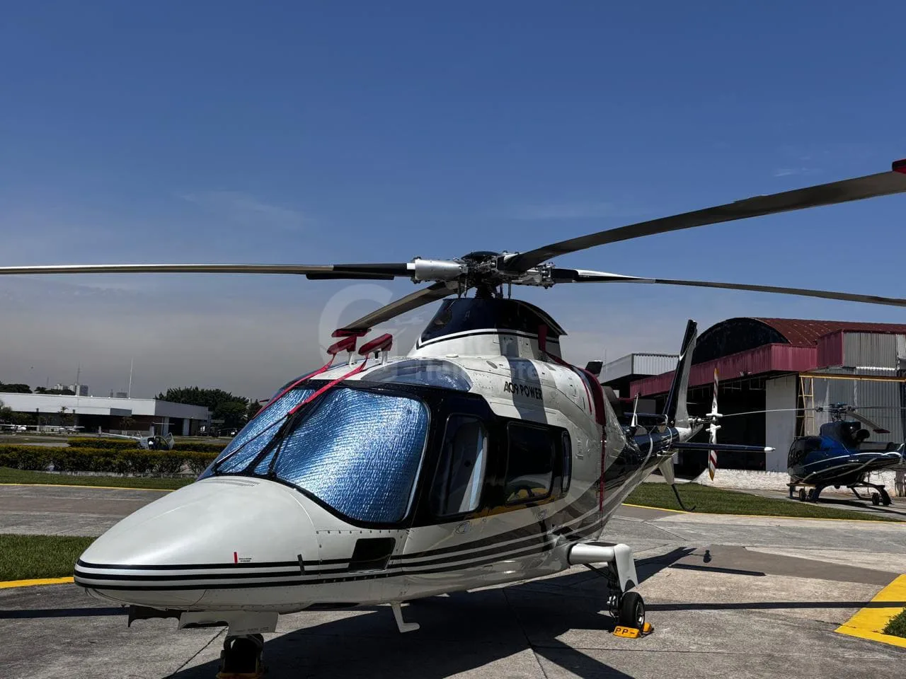 A front-side view of a white and black Agusta A109 Power helicopter with a sunshade, parked on an airfield with other aircraft in the background.