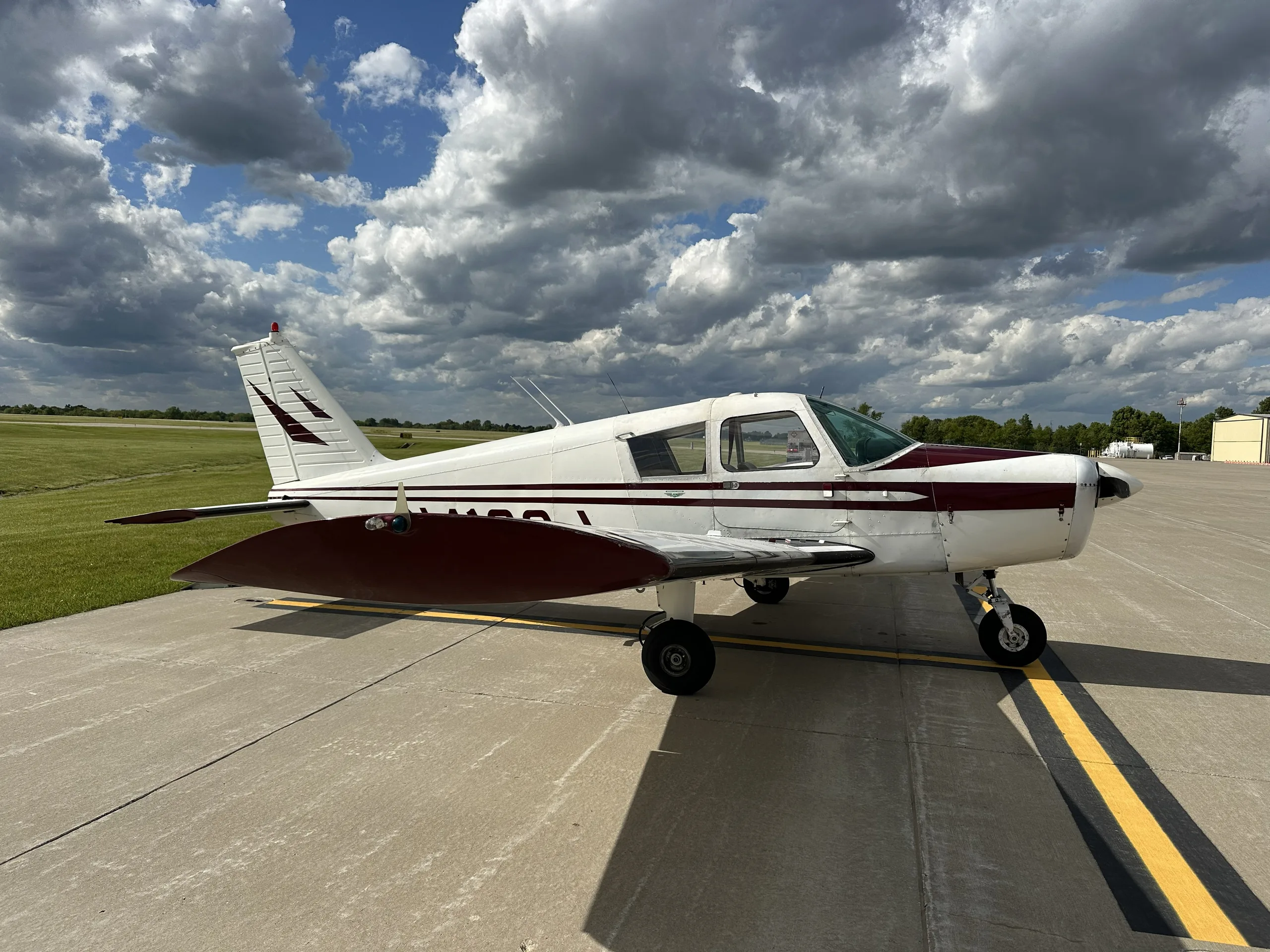 A side view of a white and maroon single-engine aircraft, likely a Piper Cherokee, parked on an asphalt ramp under a cloudy sky.