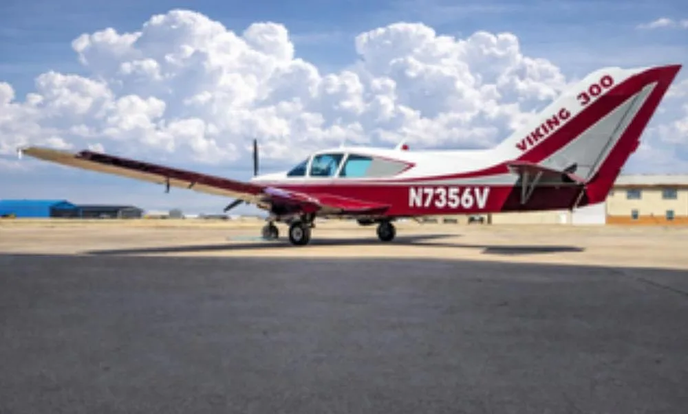 A red and white Bellanca Viking 300 (N7356V) parked on an airfield under a cloudy sky.
