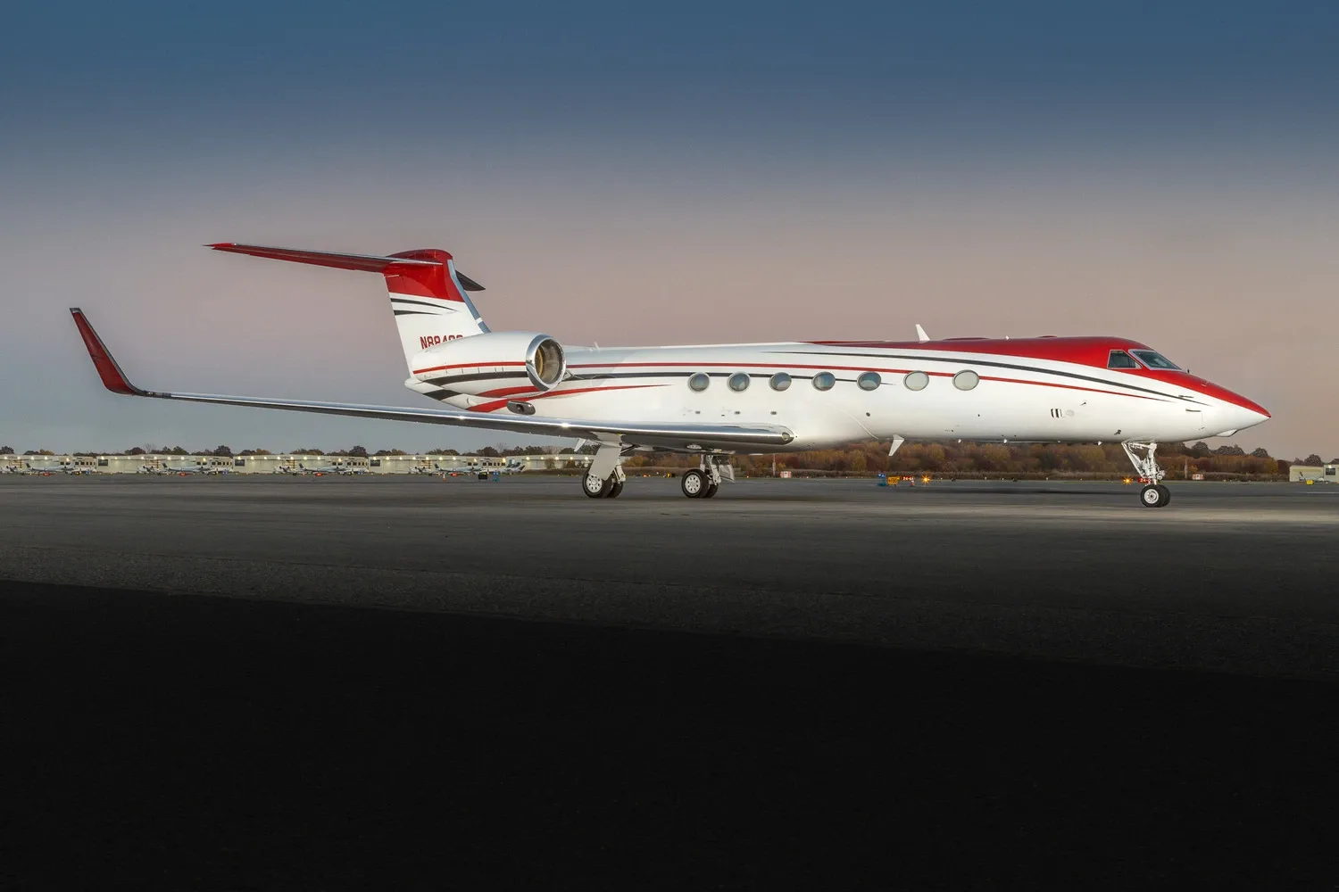 A white Gulfstream business jet with red and black stripes is parked on an airport tarmac at dusk.