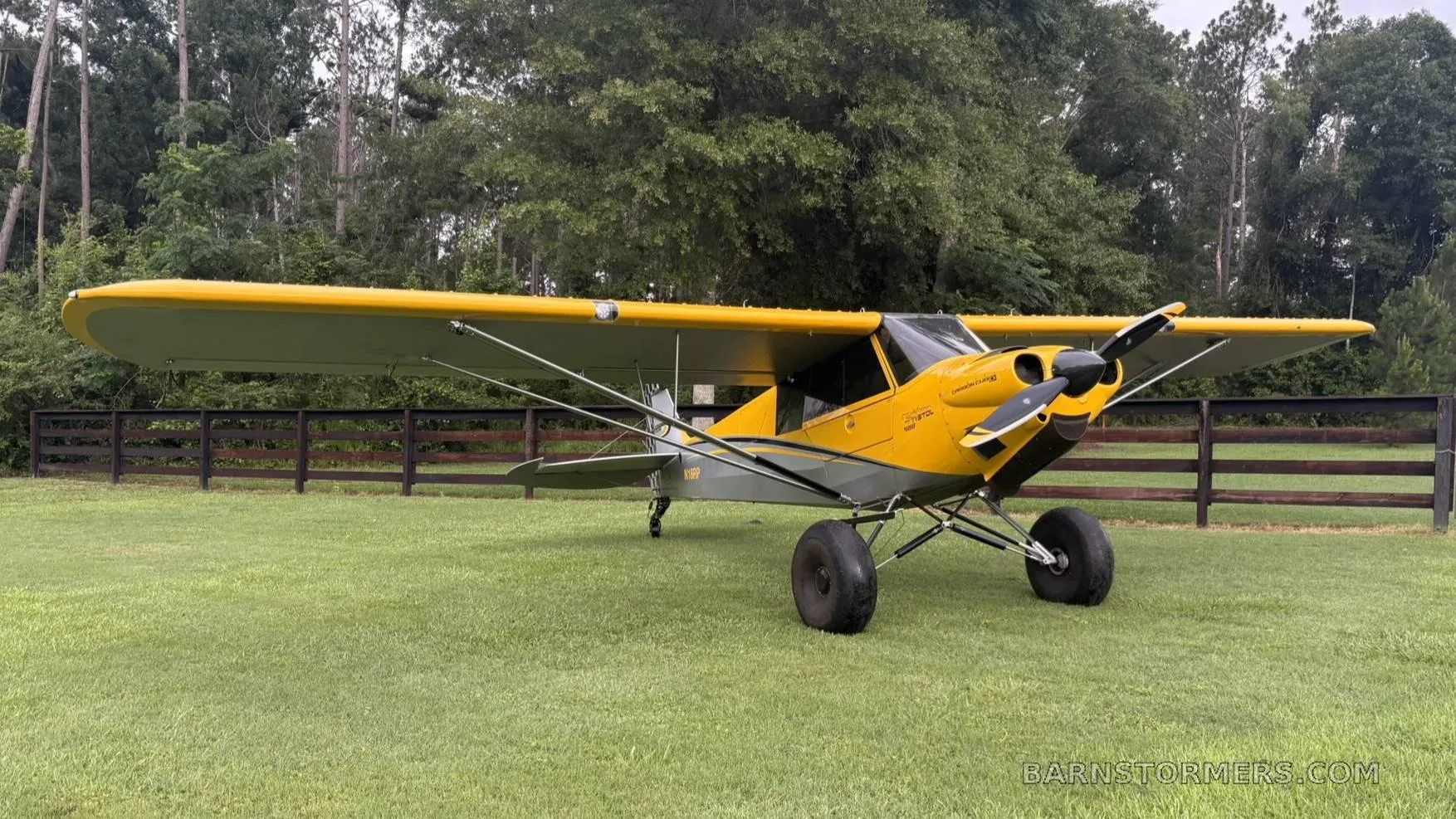 A yellow and grey CubCrafters Carbon Cub aircraft with large tundra tires, parked on a grassy field next to a wooden fence and trees.