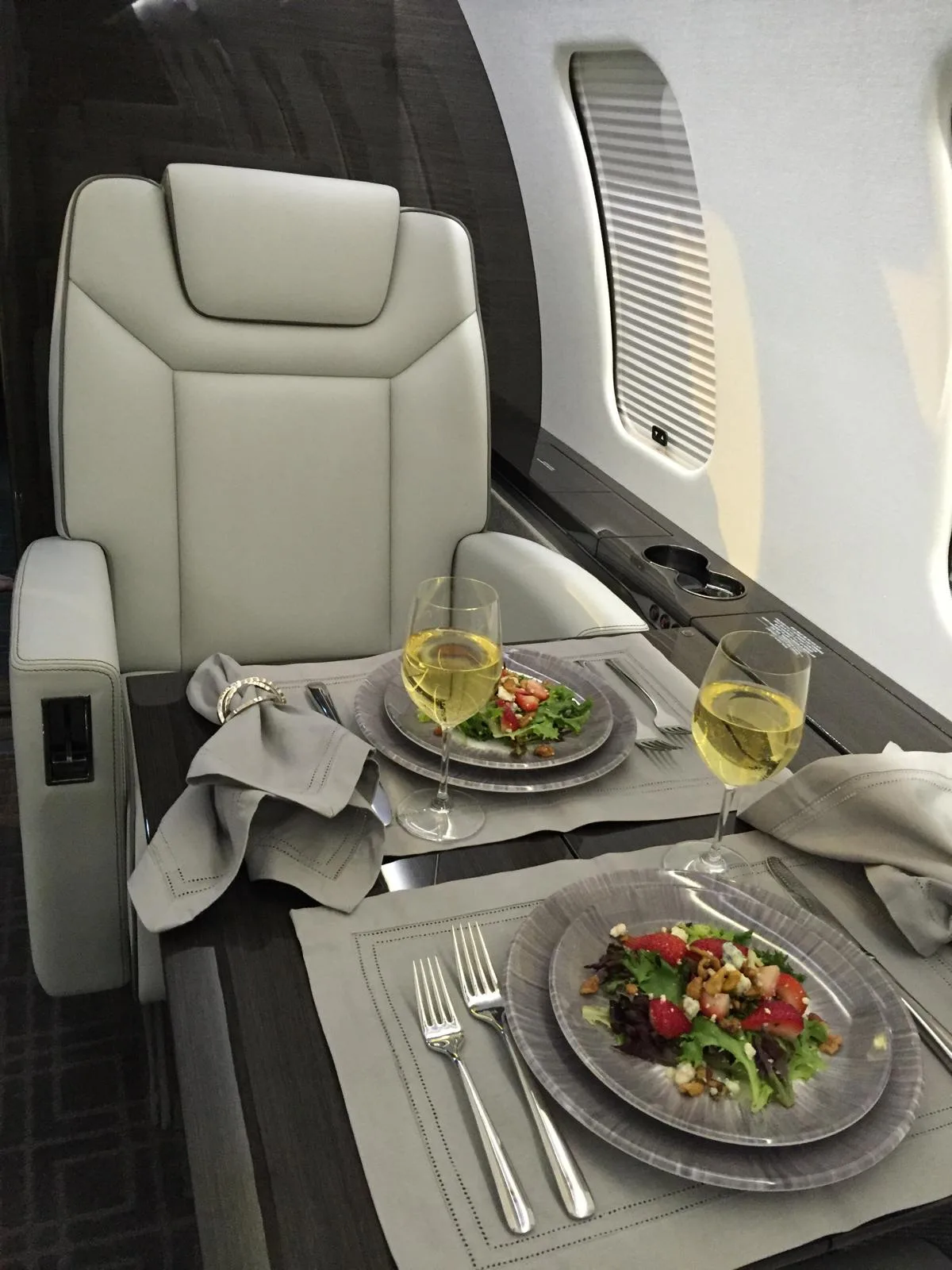 A close-up view of a private jet's interior, showing a white armchair and a table set with two salads and glasses of white wine.