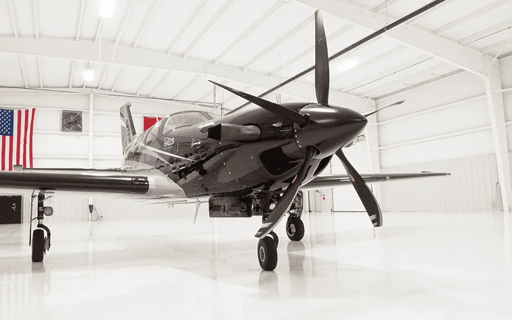 A front view of the black and silver Piper M600/SLS aircraft with its propeller, parked inside a hangar with an American flag in the background.