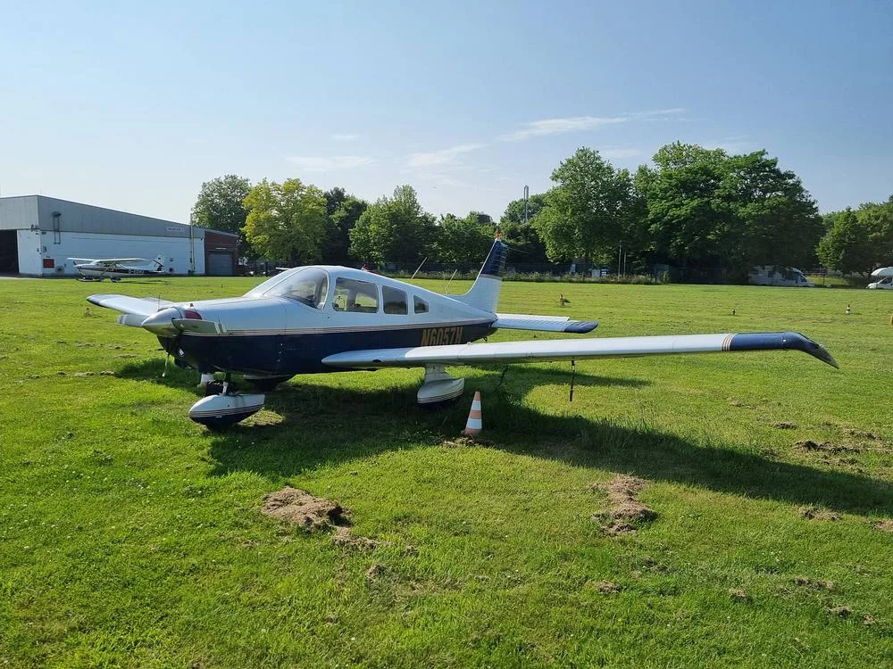 A Piper PA-28 Cherokee aircraft, registration N6057H, with blue and white paint, parked on a grassy field with hangars in the background.