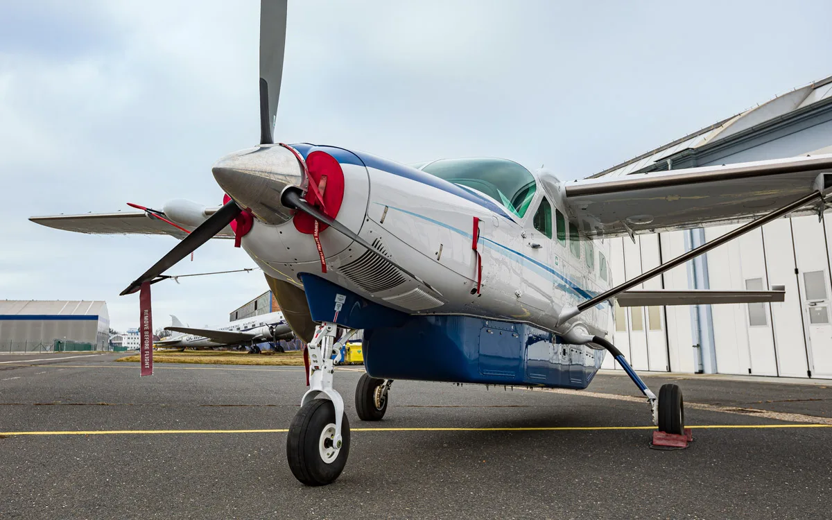 A white and blue Cessna Caravan turboprop aircraft with red 'REMOVE BEFORE FLIGHT' tags, parked on an airport apron.