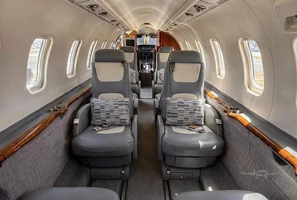 The luxurious interior of a private jet cabin features grey leather seats with patterned pillows and wood trim along the windows.