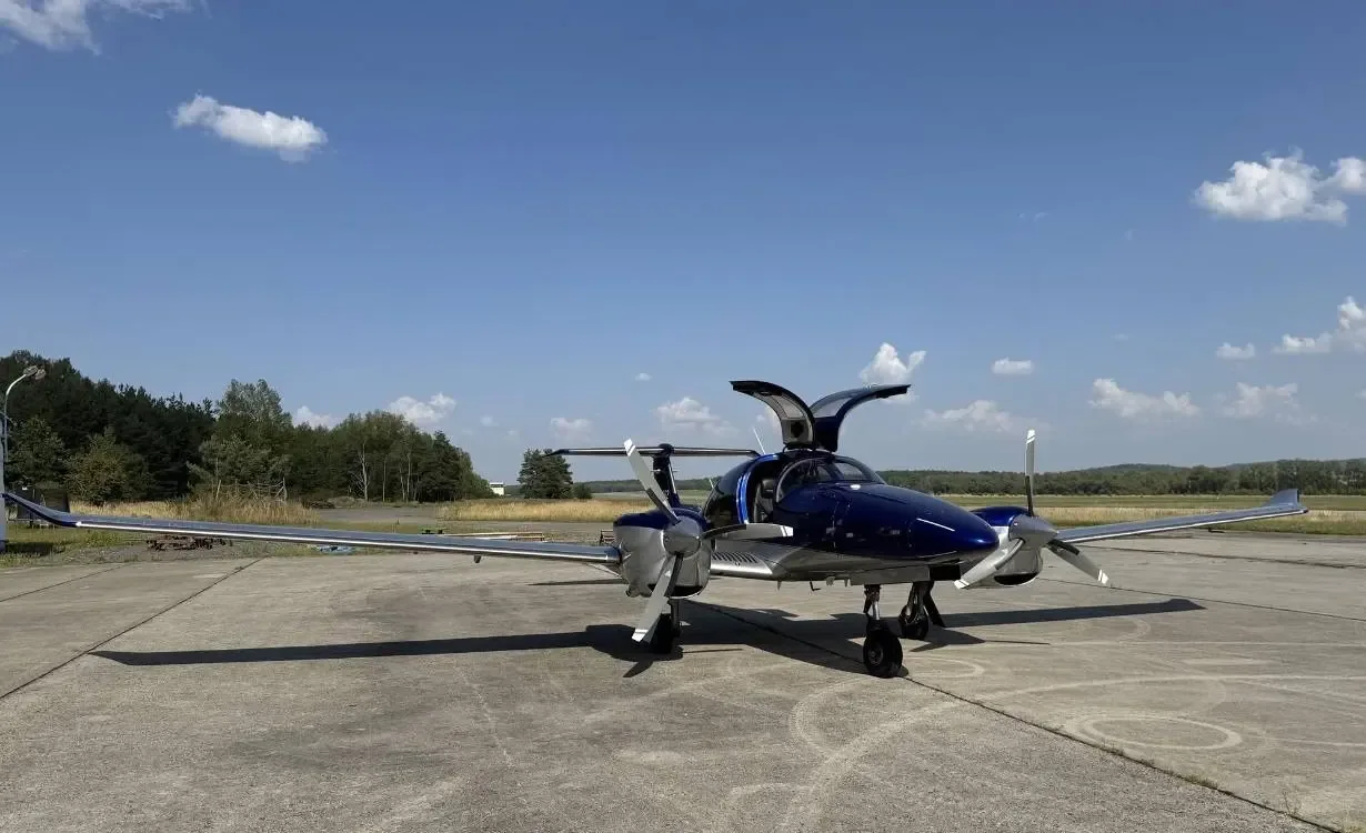 A blue and silver Diamond DA62 aircraft with its gull-wing doors open is parked on a tarmac.
