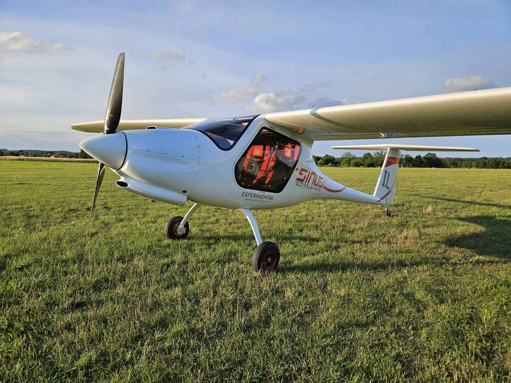 A white Pipistrel Sinus aircraft with red accents and "EXPERIMENTAL" text is parked on a grassy airfield under a partly cloudy sky.