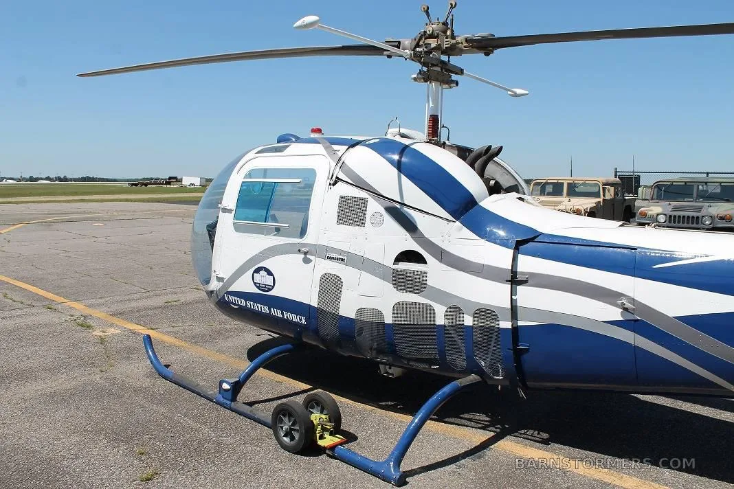 A close-up view of the front-right side of a white and blue helicopter with 'UNITED STATES AIR FORCE' text, showing its rotor and landing skids on an airfield.