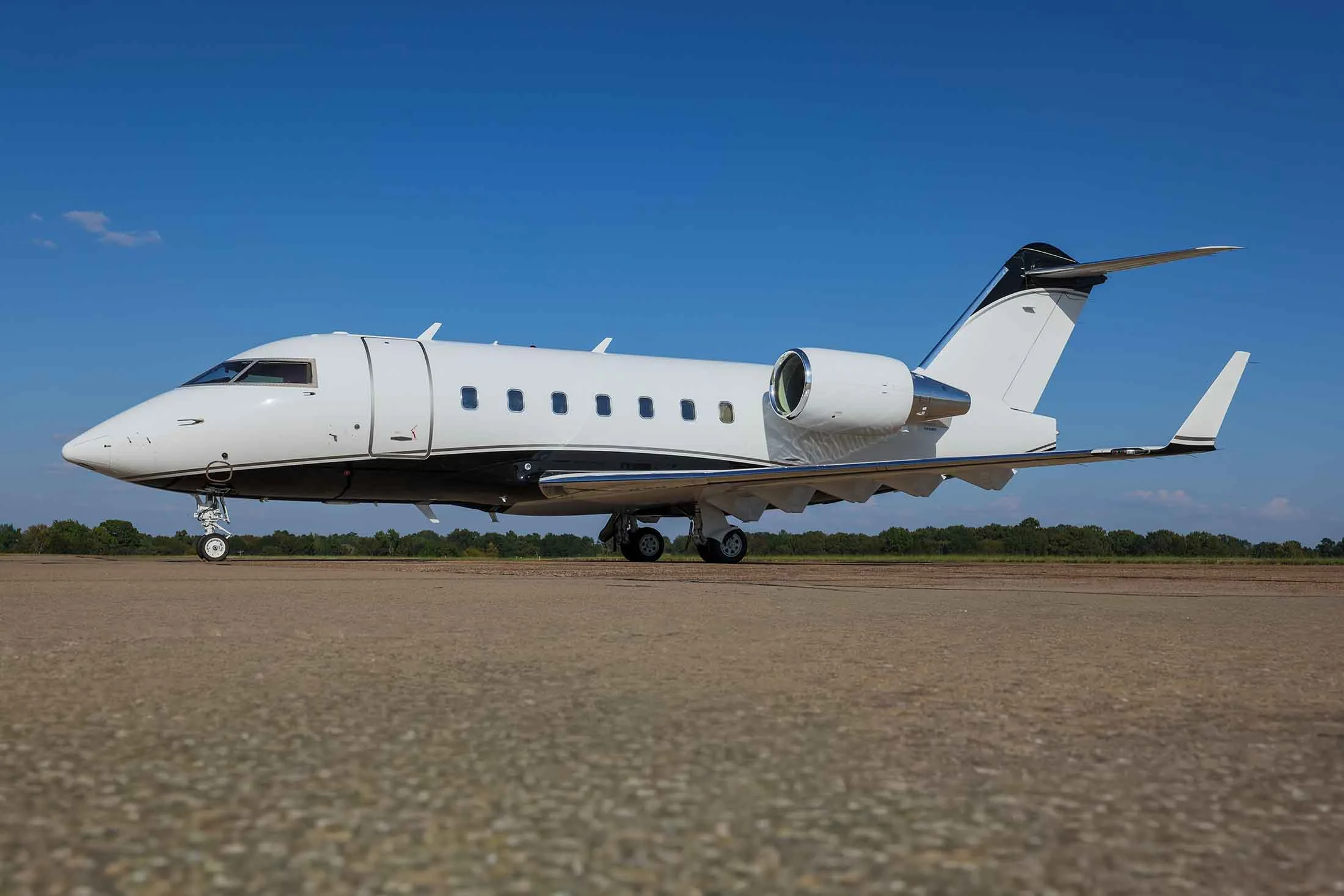 A white Bombardier Challenger business jet with black accents parked on an asphalt tarmac under a clear blue sky.