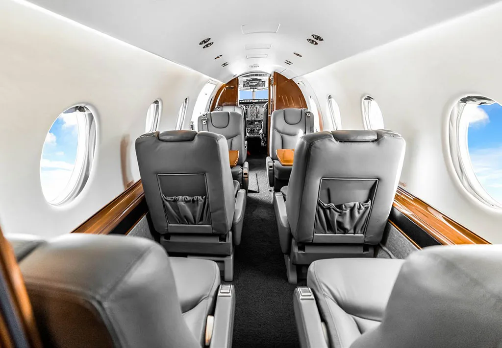 The aircraft cabin features gray leather seats and wood trim, looking forward towards the cockpit.