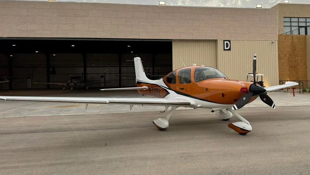 A three-quarter front view of an orange and white Cirrus SR22 aircraft parked outside a hangar.