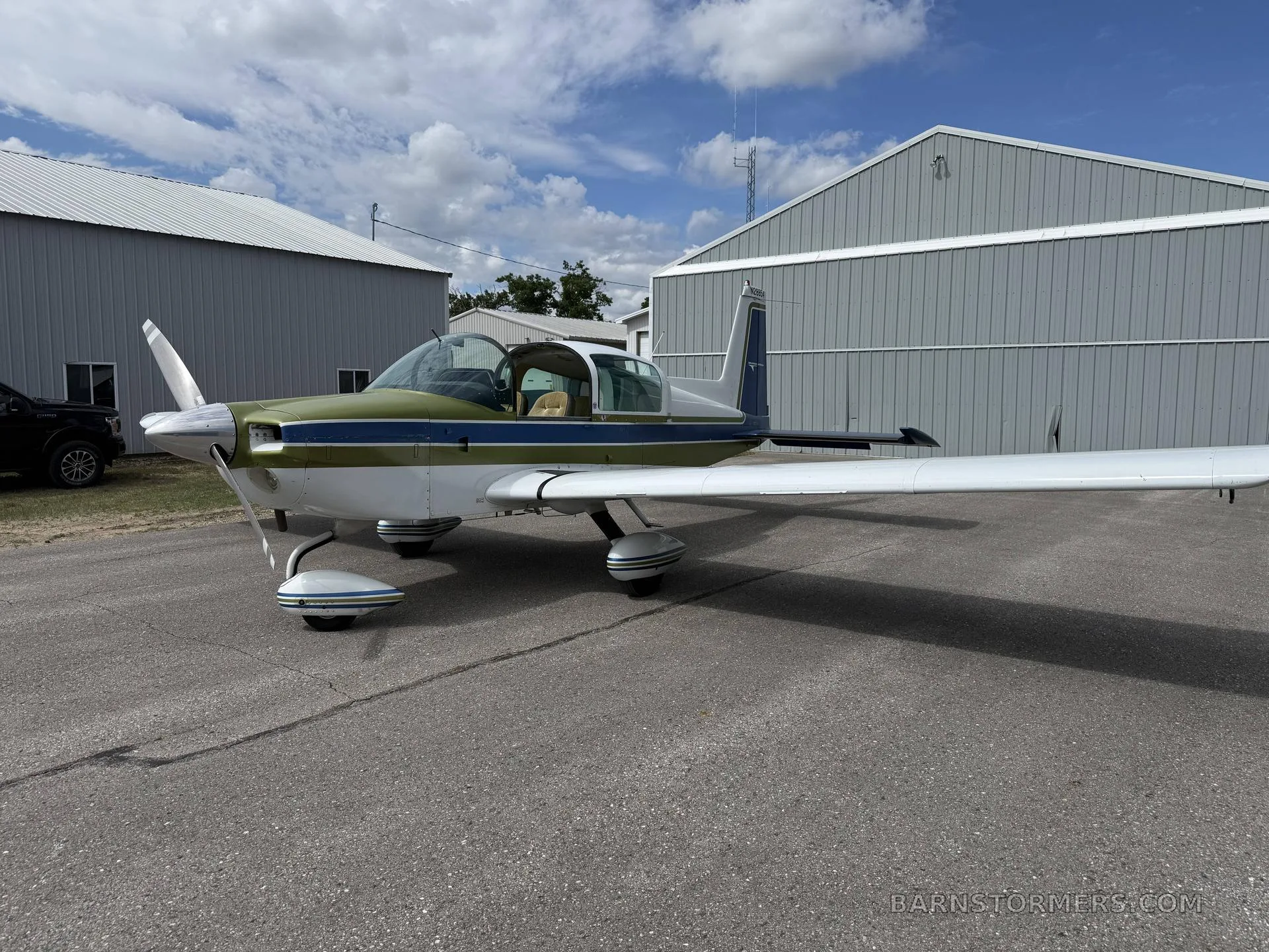 A Grumman American aircraft with green and blue stripes, parked on an asphalt ramp in front of hangars.