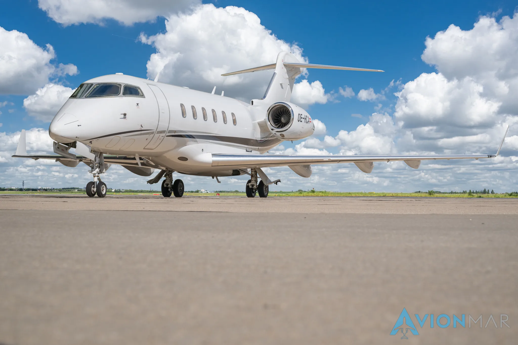 A white Bombardier Challenger 350 business jet with a black stripe on the tarmac under a blue sky with clouds, viewed from the front-right.