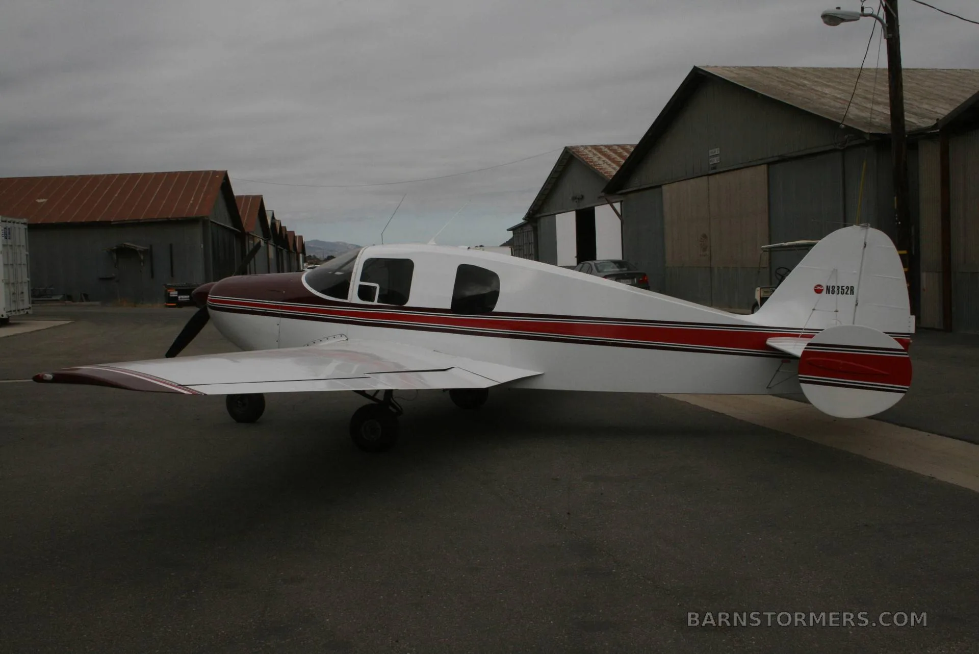 A white and maroon single-engine aircraft, tail number N8352R, parked on an asphalt apron with hangars in the background under an overcast sky.
