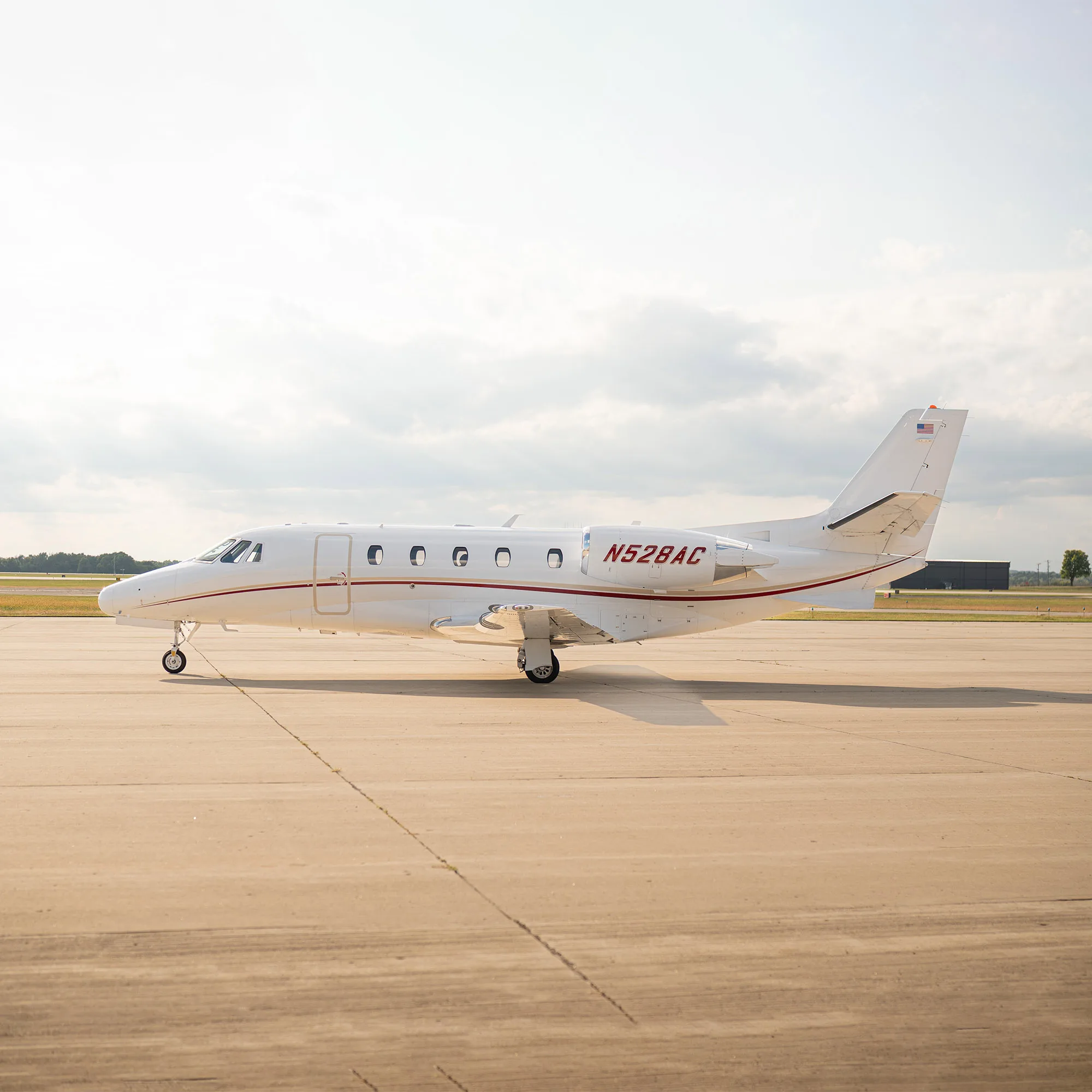 A white business jet with red stripes and registration N528AC parked on an airfield under a partly cloudy sky.