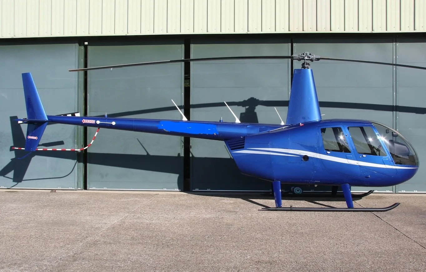 A blue Robinson R44 helicopter with white stripes parked on a tarmac in front of a hangar.