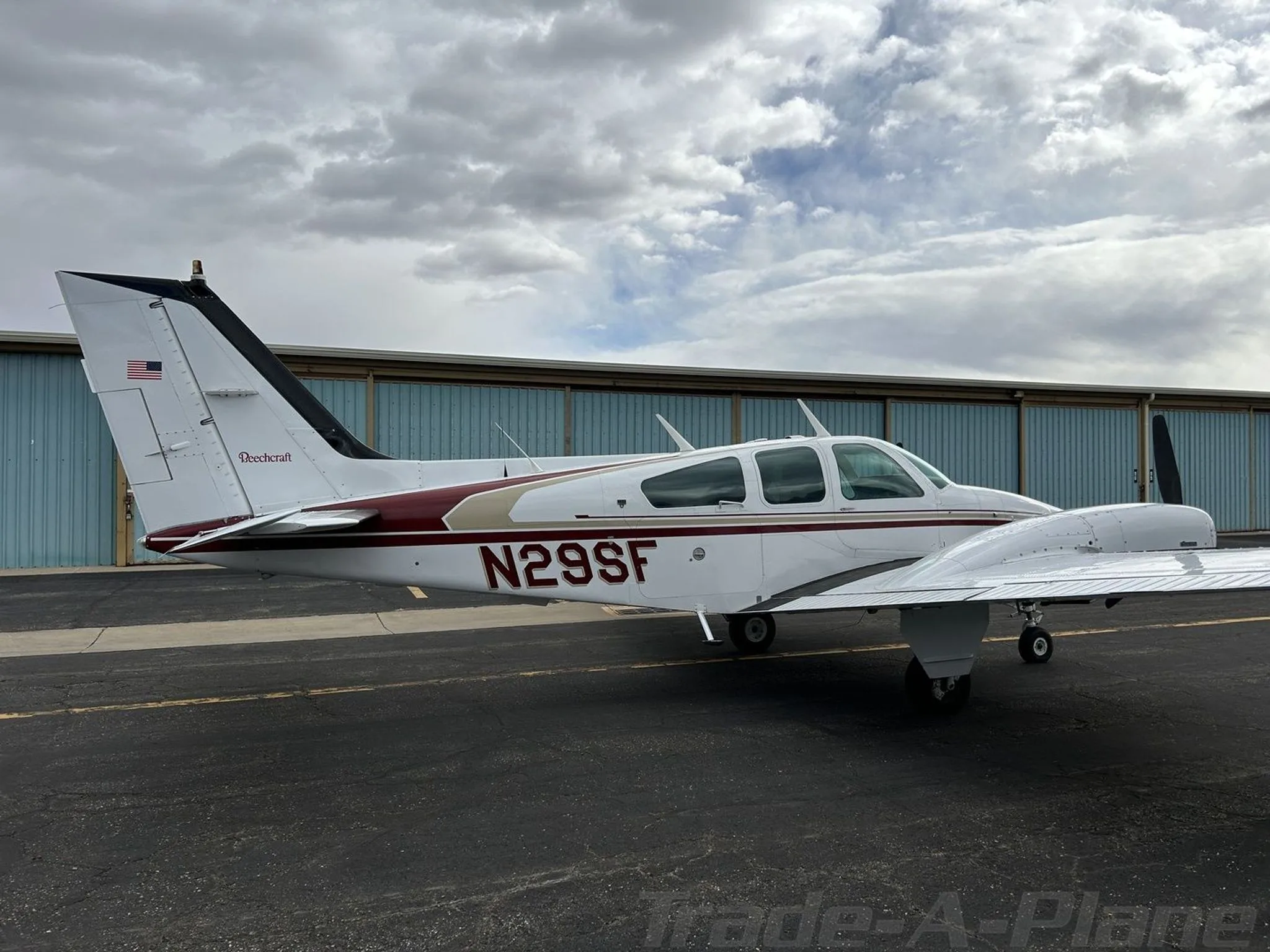 A full side view of the white and maroon Beechcraft Baron N29SF parked on an asphalt ramp with hangars in the background.