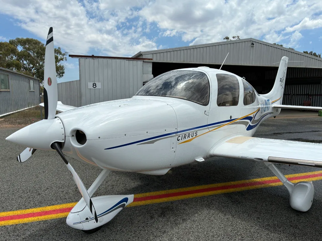 A white Cirrus SR22 aircraft with blue and gold stripes parked on an airfield in front of a hangar under a cloudy sky.