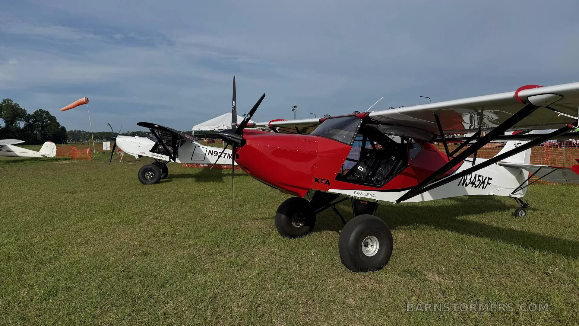 Two Kitfox aircraft, one red and white (N345KF) and one white (N927M), parked on a grassy field under a cloudy sky.
