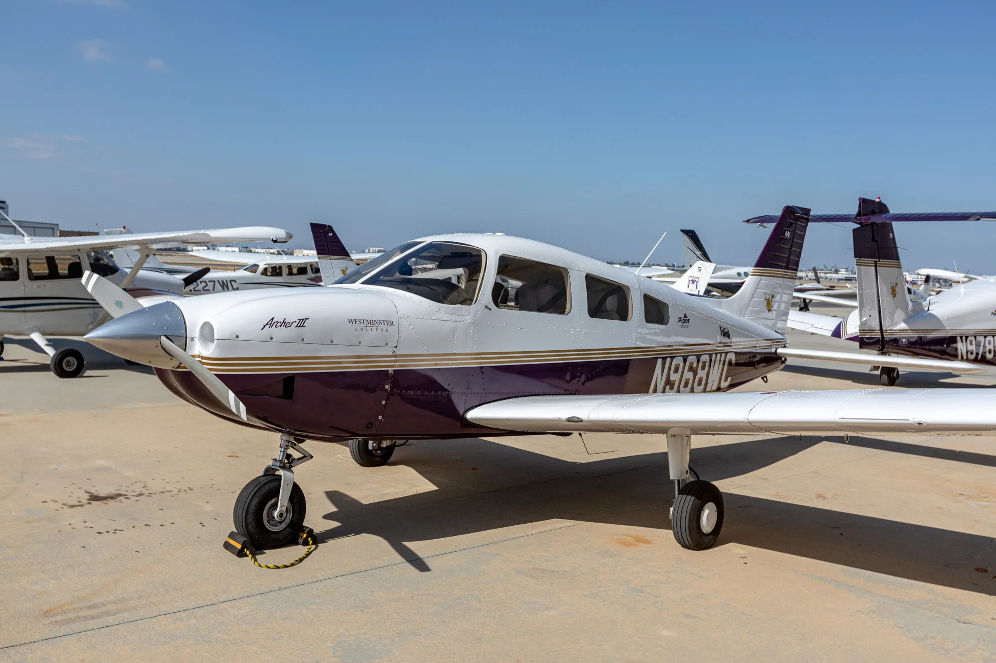 A white and purple Piper Archer III aircraft, N968WC, parked on an airfield with other planes in the background.