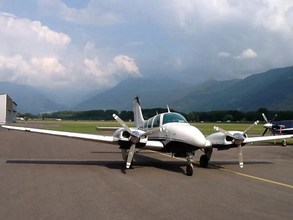 A white and black twin-engine aircraft on an airport tarmac with mountains under a cloudy sky.