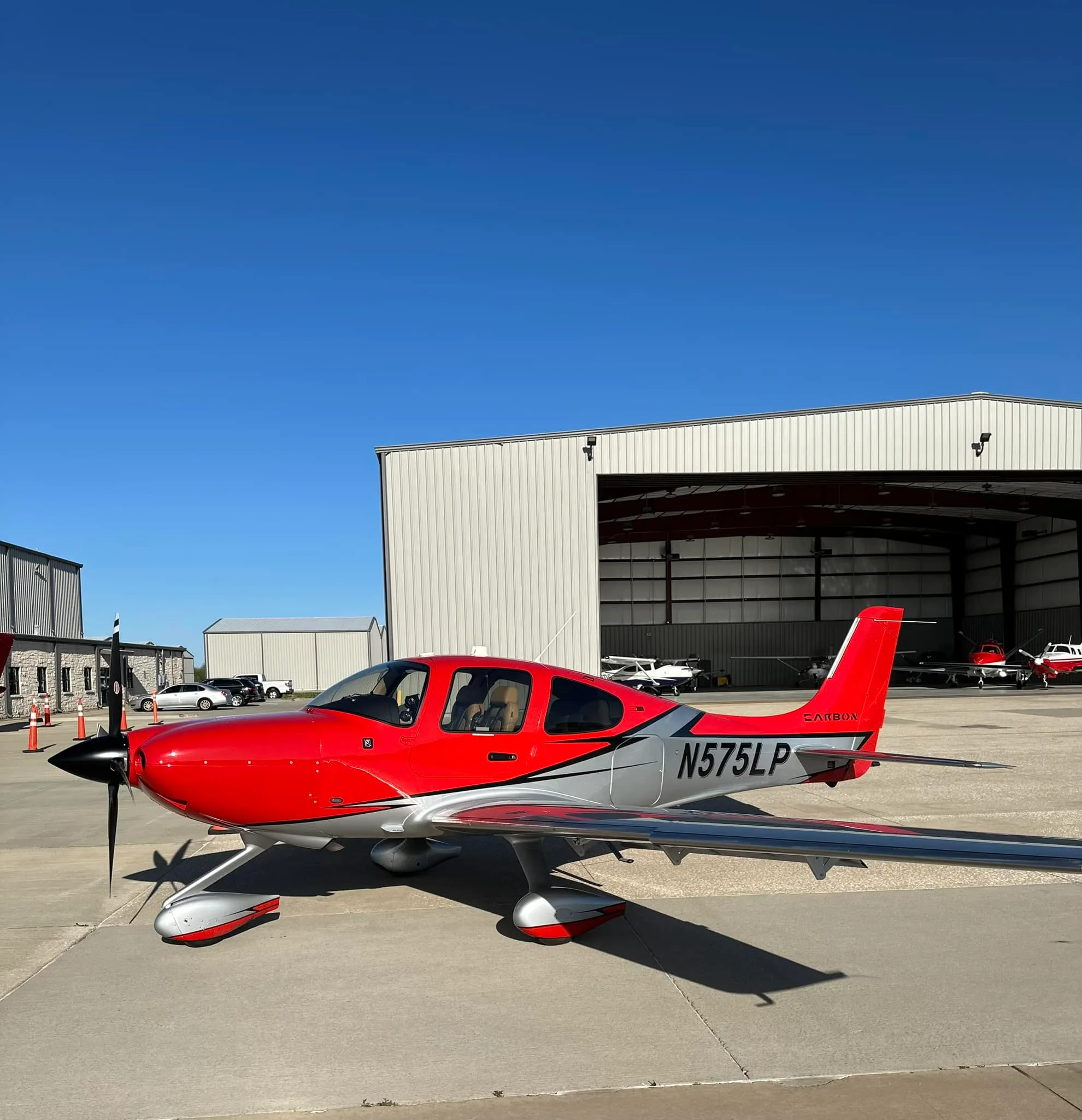 A red and silver Cirrus SR22 aircraft (N575LP) parked on an airfield tarmac in front of hangars under a clear blue sky.