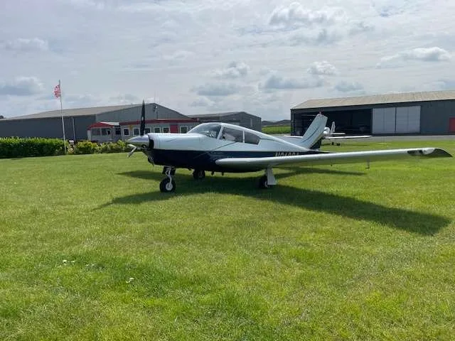 A white and blue single-engine aircraft parked on a grassy airfield with hangars in the background.