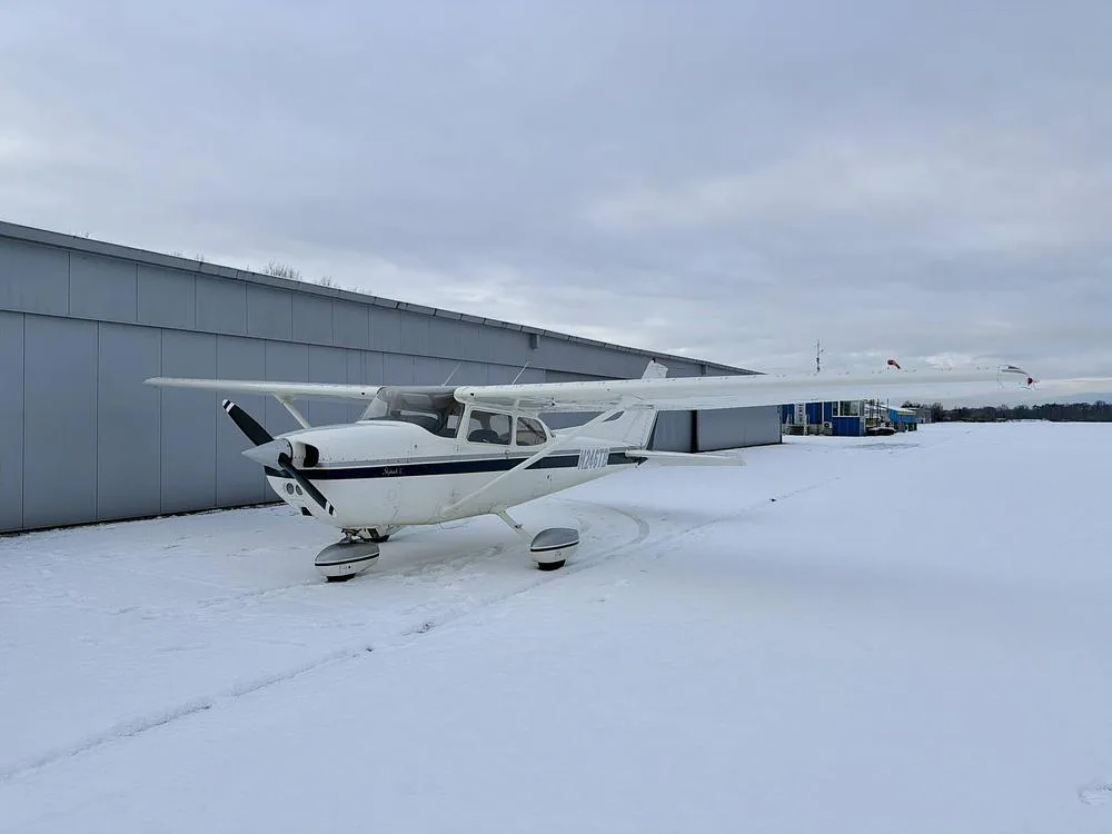 White Cessna 172 N246TB with dark blue stripes parked in the snow next to a hangar.