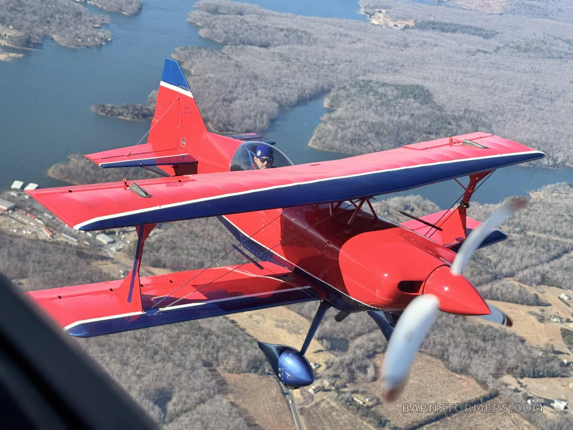 A red and blue biplane with a pilot flies over a forested landscape with lakes.