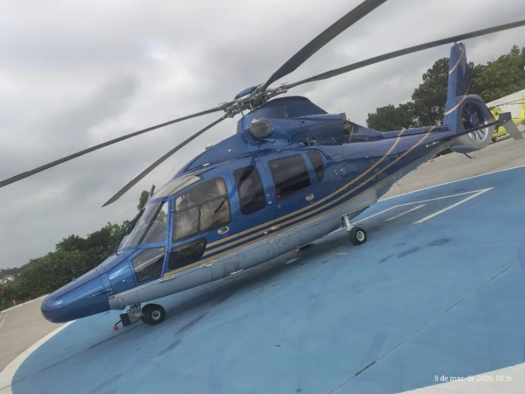 A blue and silver helicopter with gold stripes is parked on a blue helipad, viewed from the front-right.