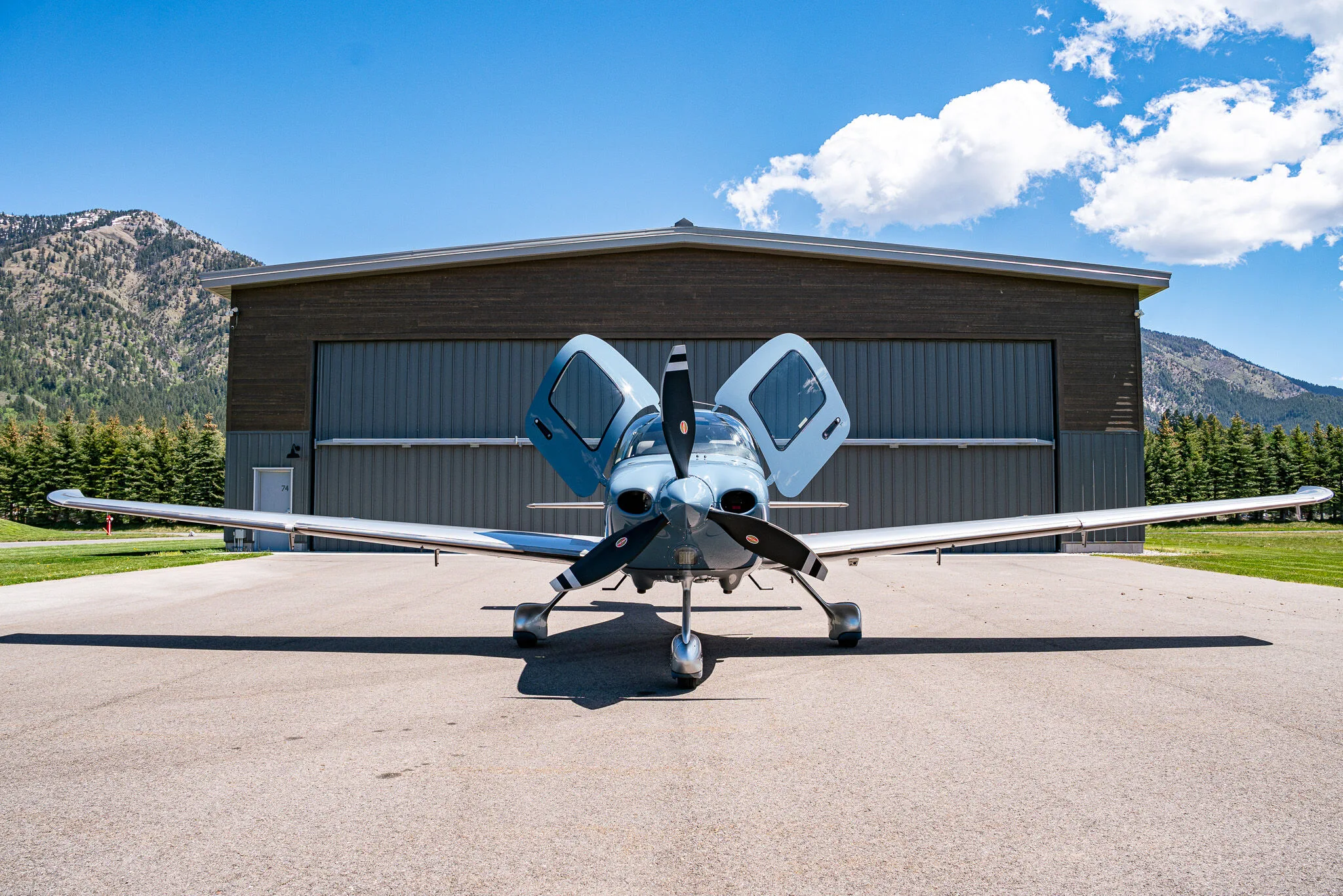 Front view of a light blue and silver Cirrus SR22T aircraft (N410HB) with its doors open, parked in front of a hangar.