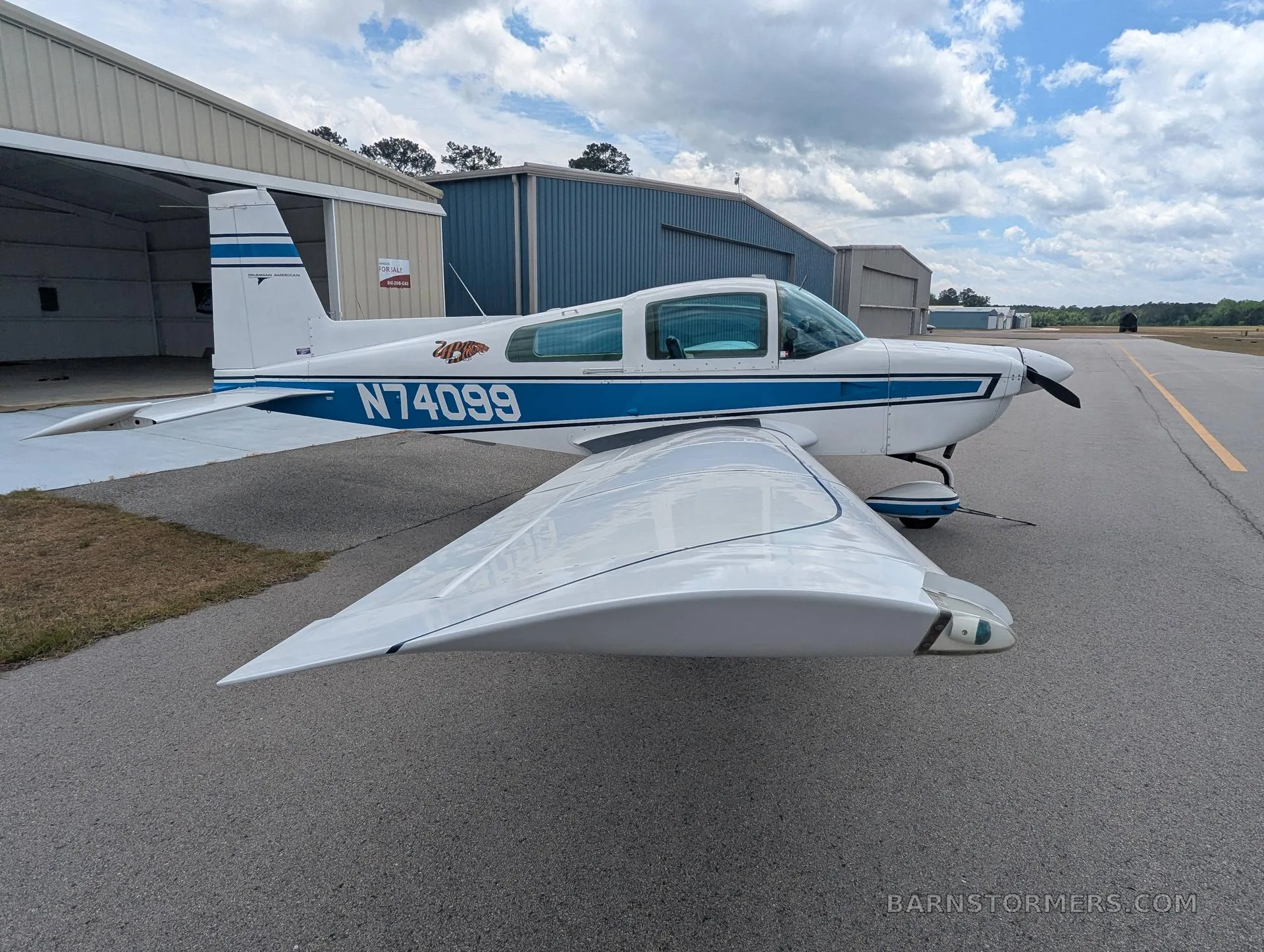 A white and blue Grumman AA-5 aircraft with registration N74099 parked on an airfield, showing its right wing and propeller.